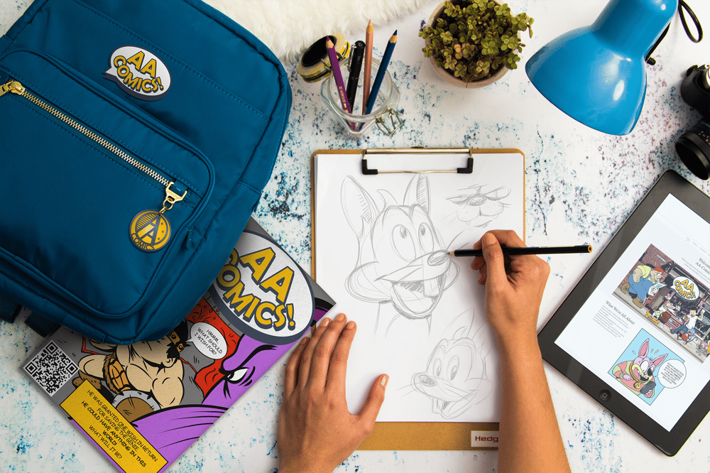 Person drawing a sketch of AA-Comics cartoons on their desk.  They have their book bag, book, tablet, pencils, camera, plant, and desk lamp on their desk. 

